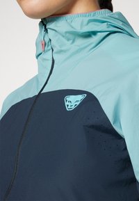 Lyseblå og marineblå teknisk jakke med glidelås i kragen, med en logo-patch og perforerte detaljer på brystet.