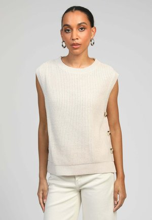 Haut sans manches en maille beige clair avec texture côtelée ; présente des accents de boutons sur le côté et un col arrondi. Associé à un jean blanc.