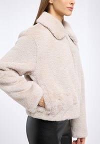 Kurtka z sztucznego futra w jasnym beżu z szerokim kołnierzem, długimi rękawami i bocznymi kieszeniami. Miękka tekstura i prosty krój.