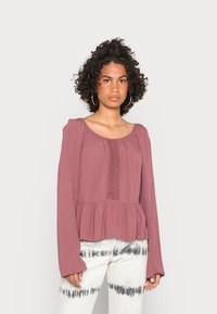 Blusa rosa poeirenta com mangas longas, decote franzido e detalhes em renda ao centro; combinada com calças brancas tie-dye.