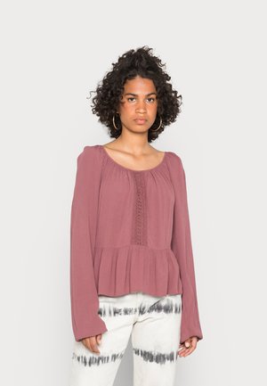 Blusa rosa poeirenta com mangas longas, decote franzido e detalhes em renda ao centro; combinada com calças brancas tie-dye.