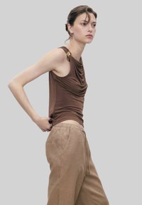 Top in tessuto drappeggiato marrone con un accento dorato sulla spalla, abbinato a pantaloni in lino beige rilassati. Texture liscia, design aderente.