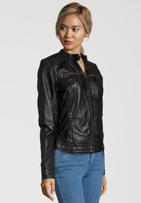 Schwarze Lederjacke mit gesteppten Schulterakzenten, Reißverschluss vorne und zwei Fronttaschen. Über blauen Jeans im Seitenprofil getragen.
