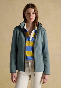 Jeune femme portant une veste polaire bleue par-dessus un polo rayé bleu et jaune et un pantalon beige clair, debout devant un fond uni.