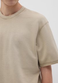 T-shirt in cotone beige con collo a giro, maniche corte e dettagli di cucitura visibili lungo le cuciture. Il tessuto ha una texture liscia.