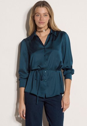 Mujer con cabello largo y rubio lleva una blusa de satén en color verde azulado oscuro con cinturón en la cintura y pantalones oscuros, de pie frente a un fondo claro y liso.