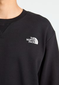 Svart genser med ribbestrikket hals; har en hvit brodert logo av "The North Face" på venstre brystområde.