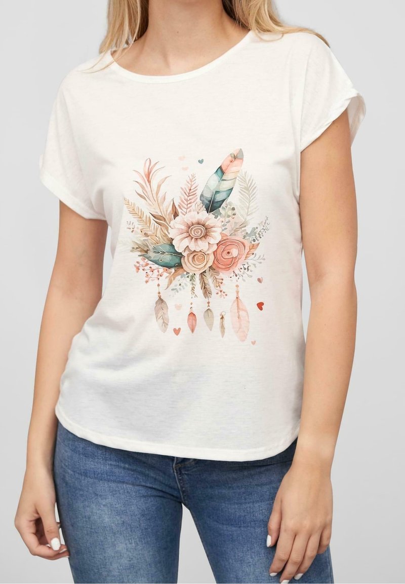 Femme portant un t-shirt blanc à manches courtes avec un motif floral et plume pastel associé à un jean bleu, debout devant un fond uni.