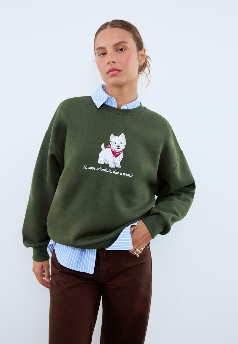 Sweat-shirt vert olive avec un chien blanc brodé portant un bandana rouge et du texte en dessous. Porté sur une chemise rayée bleu clair et un pantalon marron.