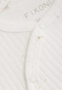 Vitt ribbat spjälsängsöverdrag med rund halsringning, framknäppning, små bruna fläckar och mjuk textur. Varumärkesetikett synlig.