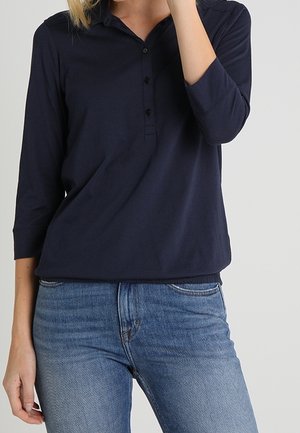 Kvinde iført en navyblå langærmet poloshirt med tre knapper og blå jeans, stående foran en ensartet lys baggrund.