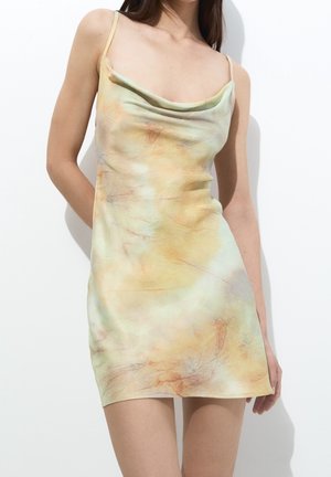 Femme portant une robe mini sans manches, tie-dye pastel avec un col drapé, debout devant un fond blanc uni.