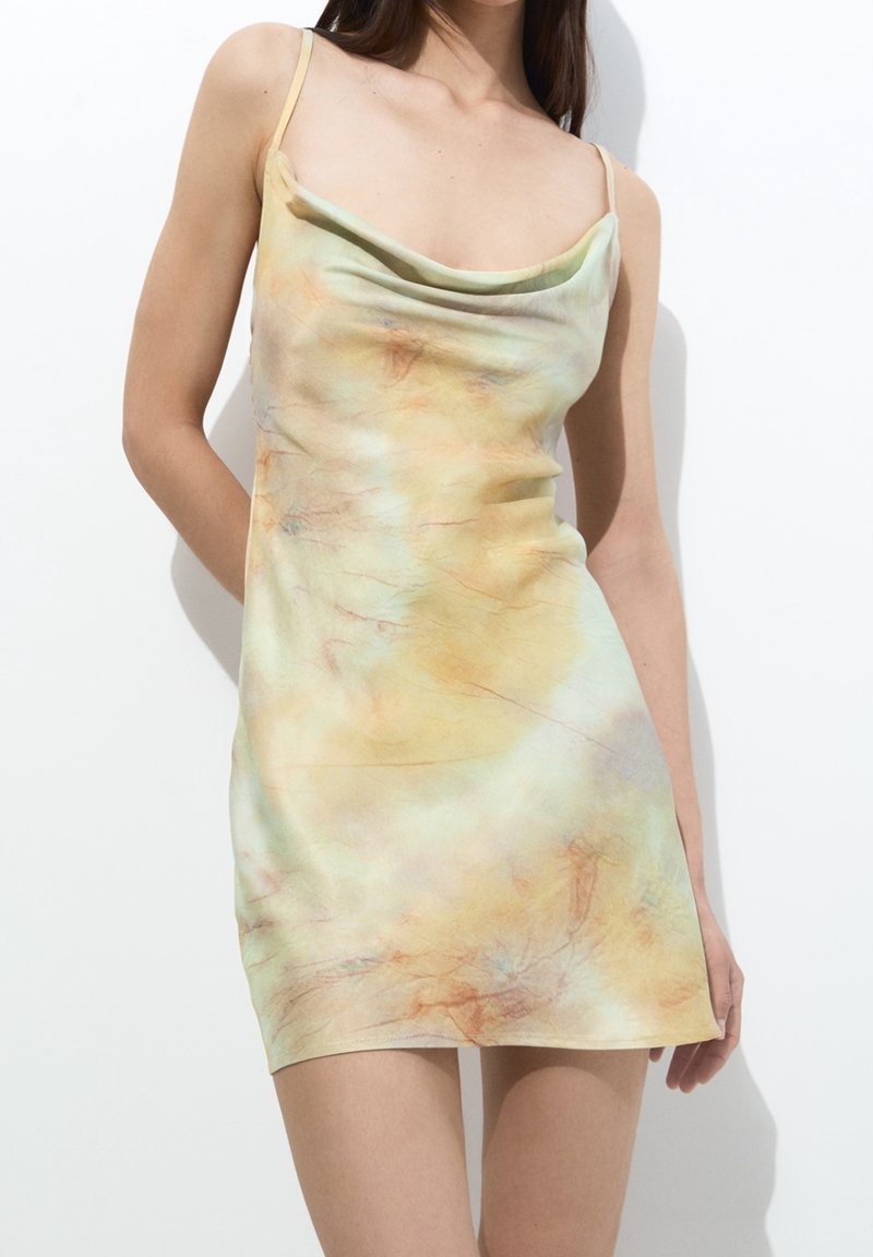 Femme portant une robe mini sans manches, tie-dye pastel avec un col drapé, debout devant un fond blanc uni.