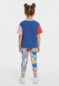 Niño con peinado de doble moño que lleva una camiseta azul de bloques de color y leggings con estampado de corazones, de pie de espaldas sobre un fondo blanco.