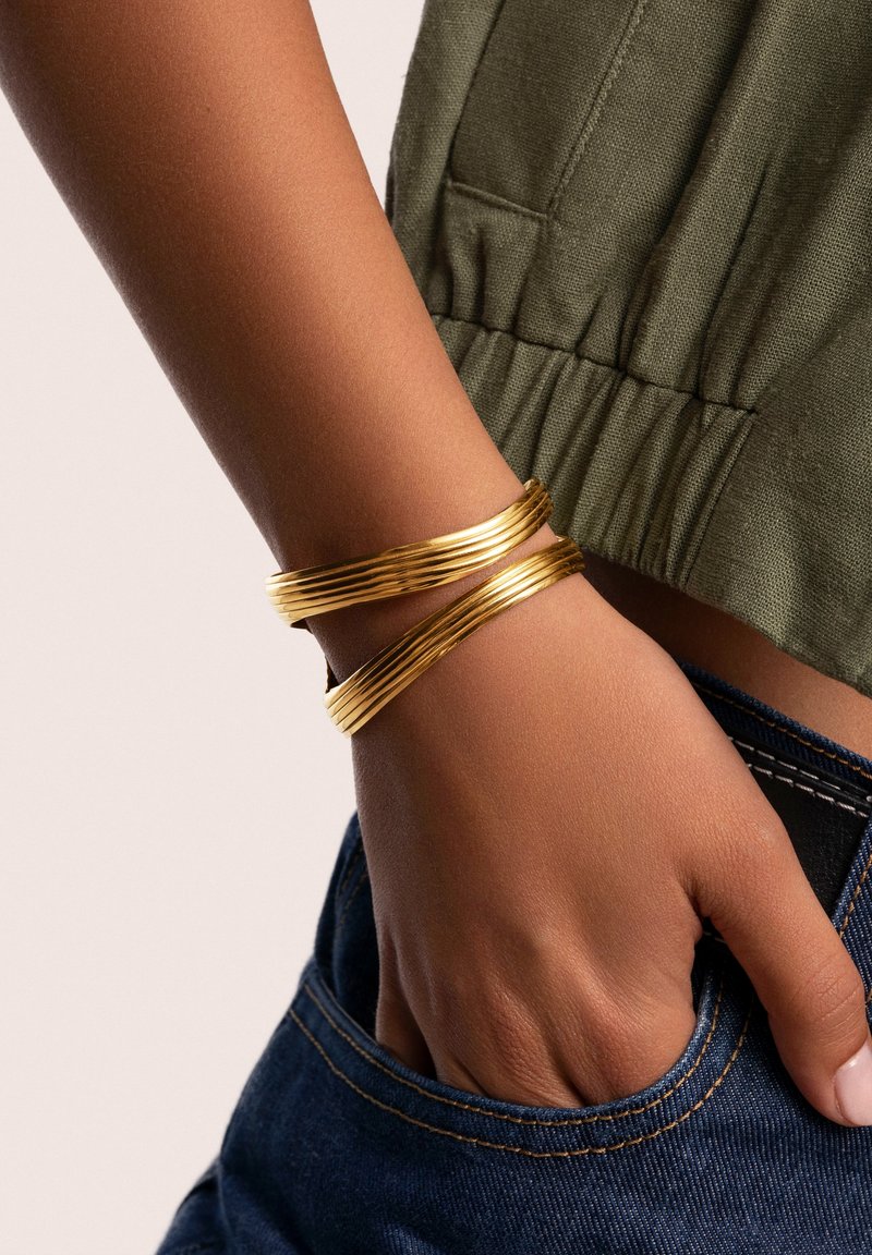 Bracciali in metallo color oro con un design testurizzato e ondulato, indossati su una mano contro una maglietta color verde oliva e jeans in denim scuro.