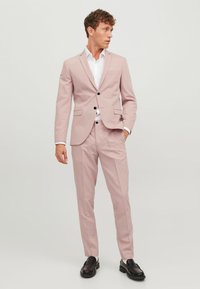 Un costume rose clair en tissu lisse, comprenant un blazer à boutonnière simple avec deux boutons et un pantalon à plis plats. Des chaussures de ville noires viennent compléter le look.