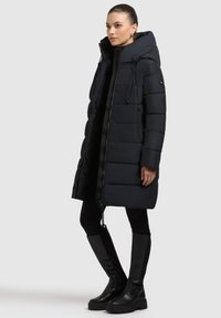 Sort polstret lang frakke med høj krave, snore og lynlås foran. Bæres over sorte leggings og kraftige sorte støvler. Glat tekstur.