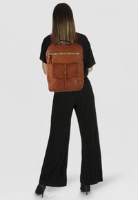 Zaino in pelle marrone con finitura testurizzata, dotato di una tasca frontale e chiusura con cerniera. Spallacci regolabili e manico superiore visibili.