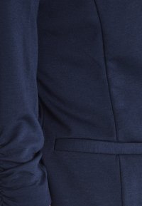 Marineblå skræddersyet blazer med en glat tekstur, synligt sidelommeklap og en let rynket ærme på venstre arm.