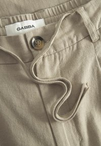 Pantalon en tissu beige avec une taille élastique, comportant une fermeture à bouton et un détail de cordon. La texture est lisse, douce et légèrement texturée.