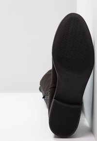Semelle d'une botte marron foncé avec un motif de bande de roulement ondulé, reposant contre un mur blanc sur une surface blanche.