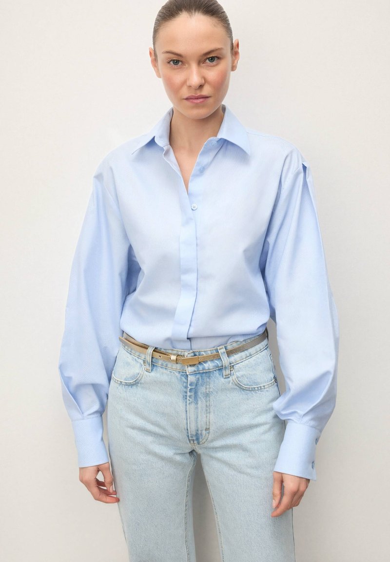 Cămașă bleu cu nasturi, guler structurat și mâneci lungi și voluminoase, asortată cu blugi deschiși la culoare și o centură bej.