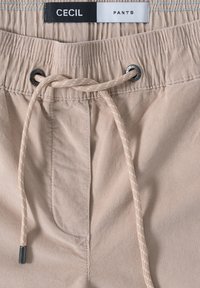 Pantalons beiges avec une taille élastique, présentant un cordon de serrage à motif beige clair et blanc, et une étiquette de marque en haut.