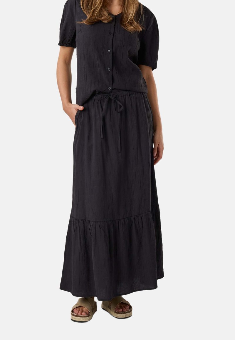 Femme portant un haut noir à manches courtes boutonné et une jupe longue assortie avec une taille à cordon, associée à des sandales compensées beige.