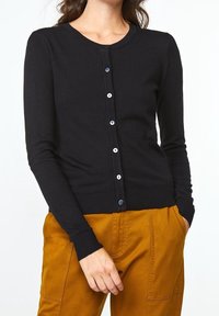 Kvinna iklädd en figursydd svart knappad cardigan med långa ärmar och senapsgula byxor, händerna i fickorna, avskuren vid nacke och midja.