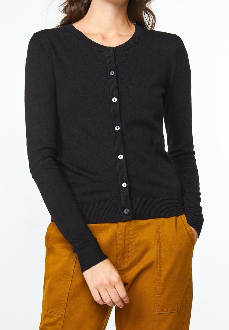 Kvinna iklädd en figursydd svart knappad cardigan med långa ärmar och senapsgula byxor, händerna i fickorna, avskuren vid nacke och midja.