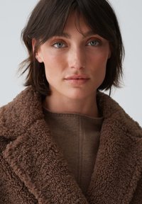 Brauner, strukturierter, flauschiger Mantel mit weitem Kragen, über einem braunen Strickpullover geschichtet. Der Stoff wirkt weich mit einer plüschigen Oberfläche.