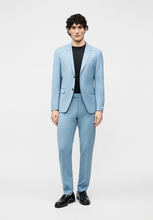 Jaunas vyras stovi, dėvėdamas šviesiai mėlyną kostiumą, juodą marškinius ir juodus klasikinės formos batus, ant vientisos šviesiai pilkos spalvos fono.