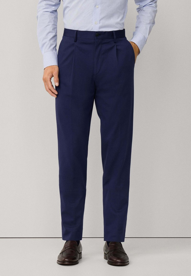 Uomo che indossa pantaloni a pieghe blu navy, camicia elegante azzurro chiaro infilata dentro, e mocassini in pelle marrone scuro, in piedi contro uno sfondo neutro.