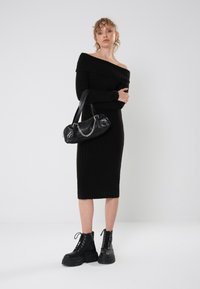 Svart ribbete off-shoulder kjole, tilpasset silhuett til midten av leggen, kombinert med en svart veske med kjedeaksenter og kraftige svarte snøresko til ankelen.
