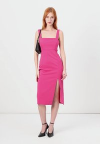 Robe rose ajustée avec un décolleté carré, une fente sur le côté et une texture lisse. Le mannequin tient un petit sac noir et porte des talons noirs.