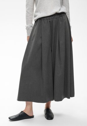 Person iført vidde mørkegrå culottes, en lysegrå bluse med lange ærmer og sorte slip-on sko mod en hvid baggrund.
