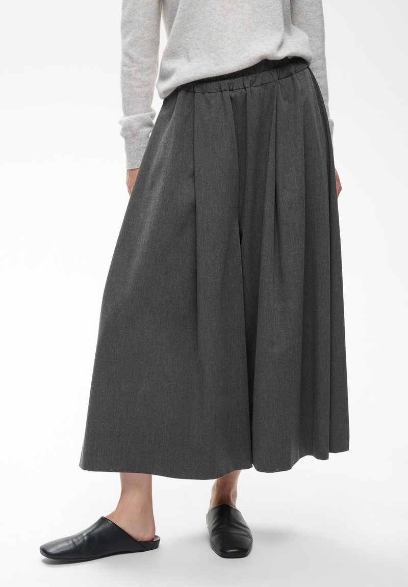 Άτομο που φοράει ανοιχτές σκούρες γκρι culottes, ανοιχτό γκρι μακρυμάνικη μπλούζα και μαύρα παπούτσια τύπου slip-on σε λευκό φόντο.
