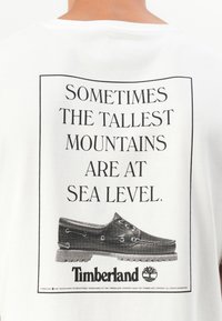 "Timberland" balta marškinėliai, su juodu grafiniu tekstu ir dėmėto rašto batų iliustracija. Apatinėje dalyje yra įmonės logotipas.