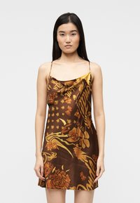 Νεαρή γυναίκα με μακριά σκούρα μαλλιά, φορώντας ένα μίνι φόρεμα από καφέ σατέν με χρυσά λουλουδένια και γραφικά σχέδια, σε απλό φόντο.