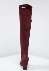 Bota de ante burdeos por encima de la rodilla con puntera redonda y tacón cuadrado. Cuenta con una cremallera lateral y una textura suave para una apariencia elegante.