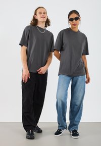 Zwei Personen stehen nebeneinander und tragen graue T-Shirts. Der Mann kombiniert seins mit schwarzen Cargo-Hosen und Sneakers; die Frau trägt hellblaue Jeans und Sonnenbrillen.