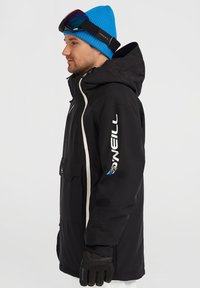 Giacca a vento nera impermeabile con cerniere e dettagli bianchi, con cappuccio, caratterizzata da un logo sul braccio. Indossata con un berretto blu e occhiali da sci.