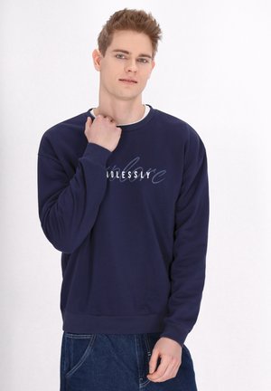 Navy blue sweatshirt with long sleeves, crew neckline, and textured “explore” print across the chest. Worn over casual denim shorts.
