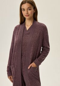 Długi rękaw, kardigan z otwartym przodem w ciemnym odcieniu mauve, wykonany z miękkiego, teksturowanego materiału. Posiada dwie przednie kieszenie oraz ściągacze przy mankietach.