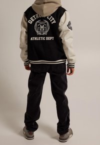 Giacca varsity nera e crema con ricamo "Detroit City Athletic Dept", abbinata a jeans scuri e sneakers grigie.