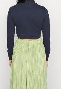 Kvinna som bär en marinblå långärmad crop top tillsammans med en högmidjad, ljusgrön plisserad kjol, sedd bakifrån mot en enkel bakgrund.