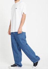 Homme portant un jean bleu ample, un T-shirt blanc oversize et des baskets basses noires sur un fond blanc uni.
