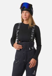 Zwarte skibroeken met witte merkbanden, voorzien van een hoge tailleband, ritssluitingen en een gladde matte textuur. Gecombineerd met een muts en skibril.