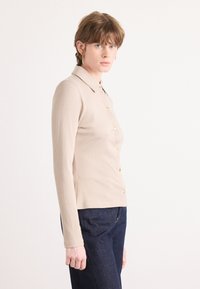 Langärmliges, hellbeiges, geripptes Polohemd mit Kragen und goldenen Knopfdetails, kombiniert mit dunkelblauen Jeans. Weiche Textur.