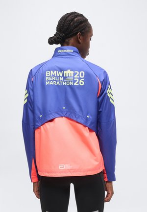 Personne aux cheveux tressés porte une veste bleue et corail étiquetée "BMW Berlin Marathon 2026" et "#BERLINLEGEND" sur le col.